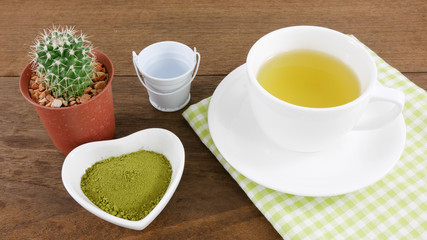 The Japanese matcha green tea powder on ceramic heart shaped bowl and cup of hot green tea with cotton fabric on wooden planks.