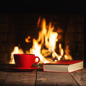 Red cup of coffee or tea and old book on wooden table near  fire