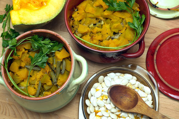 Still-life with homemade fried pumpkin with carrot and green beans in color clay pots with raw squash and seeds in a plate on kitchen table. Top view close-up