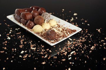 Assorted chocolate candies on white plate with flakes over black background