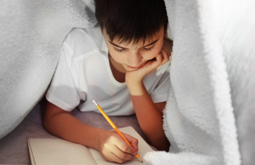 Little boy writing with pencil under the blanket