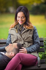 Happy young woman sitting on a bench in a park with her boyfriend lying on her lap