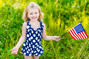 Smiling little girl with long curly blond hair holding american flag and waving it, outdoor portrait on sunny day in summer park. Independence Day, Flag Day concept