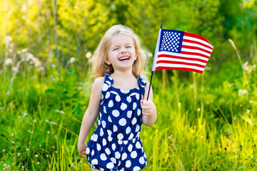 Adorable laughing little girl with long curly blond hair holding american flag and waving it on sunny day in summer park. Independence Day, Flag Day concept