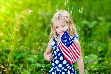 Cute smiling little girl with long curly blond hair holding an american flag on sunny day in summer park. Independence Day, Flag Day concept