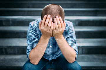 Outdoor portrait of sad young man covering his face with hands sitting on stairs. Selective focus...