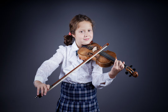 Beautiful little girl with curly hair wearing blue shirt and checked skirt playing the old violin, studio portrait  on dark background