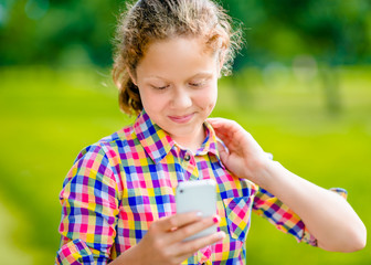 Adorable smiling teenage girl in casual clothes with smartphone in her hand, looking at screen, reading a message, using social network on sunny day in summer park