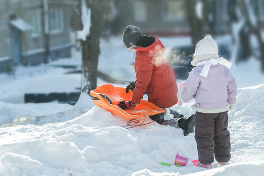 Playing sibling children preparing for winter riding downhill on orange plastic snow slider outdoors