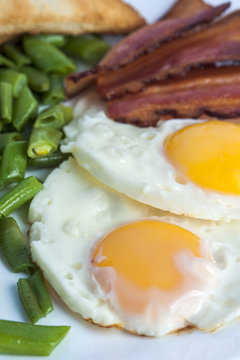 Fried eggs, bacon, green beans and toasts on light background. English breakfast. Vertical view. Focus on eggs.