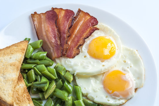 Fried eggs, bacon, green beans and toasts on white plate on light background. English breakfast. Horizontal view.
