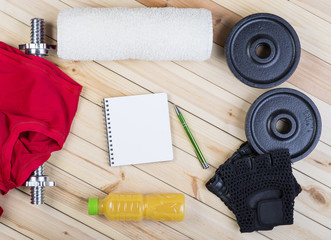 Sport Equipment. Barbell, Weights, Red Shirt, Orange Juice, Towel, Gloves And Notebook To Workout Plan On Wooden Table. Sport Fitness Background
