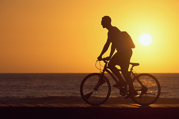 Fototapeta na wymiar Silhouette of young man cyclist on sunset sky with Sun on water texture with ripples