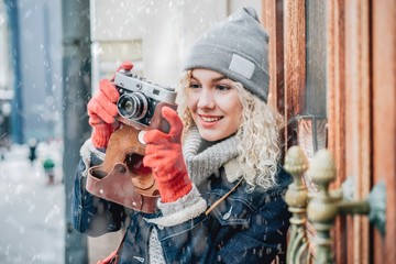 Young blond curly female in warm clothers with old film camera shooting a photo on the background of wooden doors with handles, winter, snow