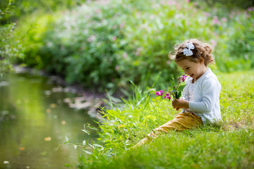 Sweet, happy little girl sitting on a grass in a park at a spring stream with flower in hand....