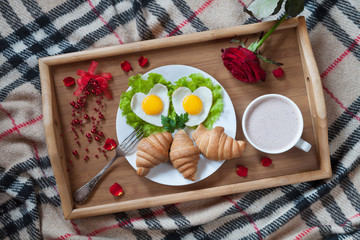 Romantic breakfast in bed with heart-shaped eggs, jam toasts, croissants, rose flower and petals