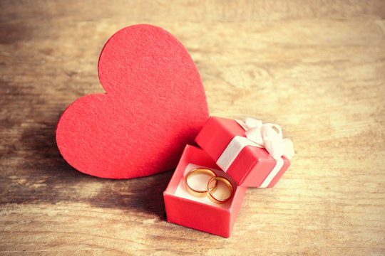Red heart and wedding rings in a box on wooden background closeup