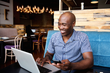 Smiling man with a mobile phone sitting at cafe using laptop
