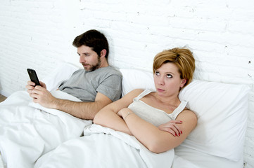 young couple in bed unsatisfied wife bored frustrated and angry while internet addict husband is using mobile phone  social network obsession