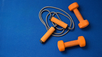 Pair of orange dumbbells and jumping rope on blue yoga mat background