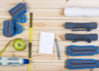 Sport Equipment. Dumbbells,  Ankle Weights, Wrist Weights, Towel, Tape Measure, Apple, Bottle Of Water And Notebook To Workout Plan On Boards. Sport Fitness Background