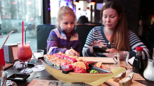 Mother and young daughter sitting in asian restaurant with big set of shushi and rolls, woman photographing food with cellphone
