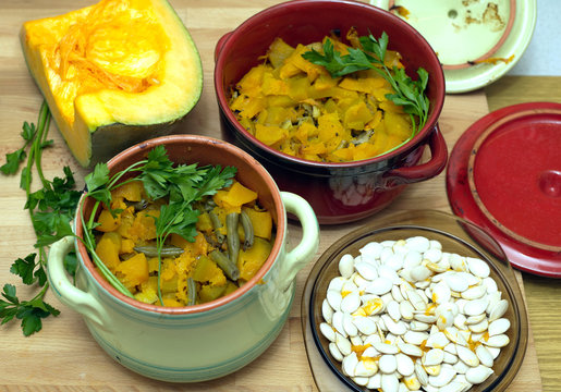 Still-life with homemade fried pumpkin with carrot and green beans in color clay pots with raw squash and seeds in a plate on kitchen table. Top view close-up