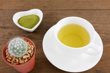 The Japanese matcha green tea powder on ceramic heart shaped bowl and cup of hot green tea and little cactus in plant pot on wooden planks.