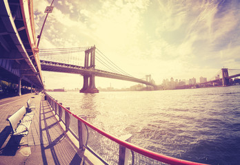 Vintage stylized fisheye lens photo of bench at East River bank and Manhattan Bridge in distance against sun.