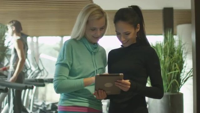 Two beautiful athletic women are using a tablet next to treadmills in a sport gym. Shot on RED Cinema Camera.