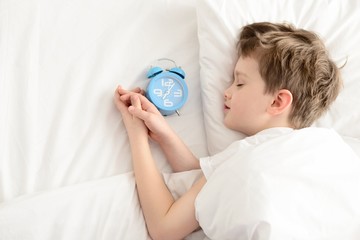 Top view of little boy sleeping in white bed with alarm clock near his head.