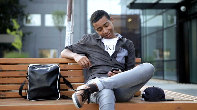 Young guy is chatting a message for his classmates in his mobile phone . He is sitting on a beanch in front of his university