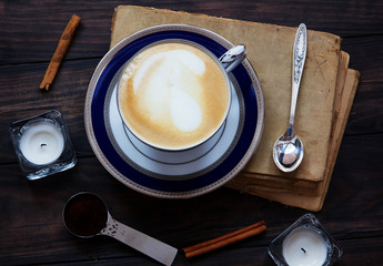 Coffee composition with old book, white tea candles, silver spoon, ground coffee and cinnamon on dark wooden background.