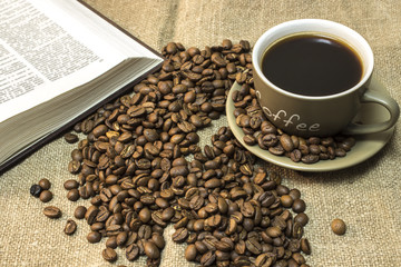 A cup of coffee with scattered coffee beans and open book are  stands on a piece of burlap