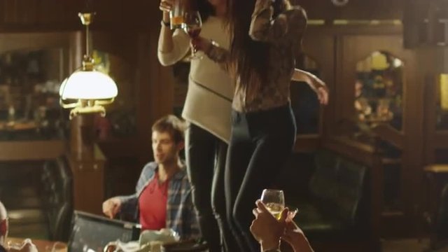 Two girls are dancing with drinks on a table while everybody are having a good time together at a bar. Shot on RED Cinema Camera.