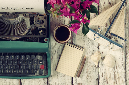 image of vintage typewriter with phrase "Follow your dreams", blank notebook, cup of coffee and old sailboat on wooden table