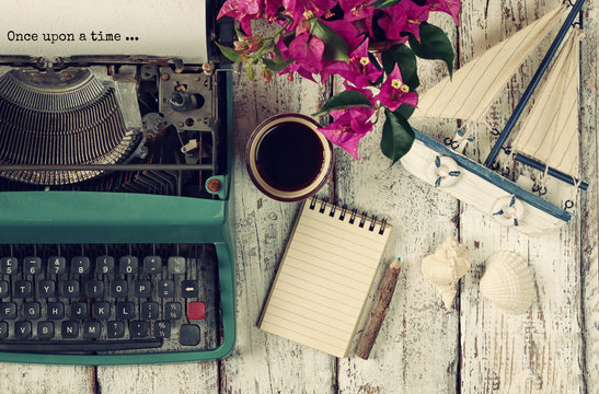 image of vintage typewriter with phrase "once upon a time", blank notebook, cup of coffee and old sailboat on wooden table
