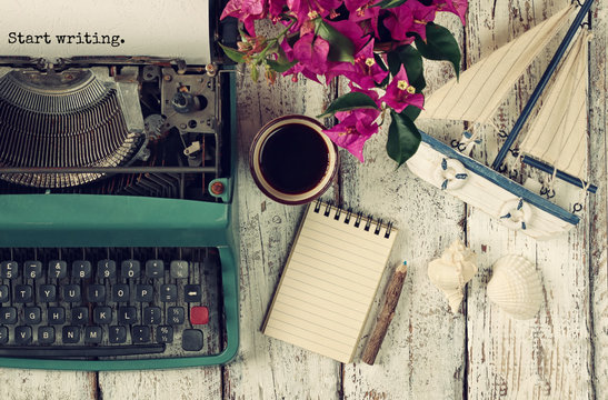 image of vintage typewriter with phrase "Start writing", blank notebook, cup of coffee and old sailboat on wooden table