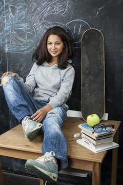 young cute teenage girl in classroom at blackboard seating on table smiling