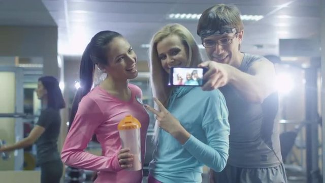 Two attractive fit sporty girls and a man are making selfie pictures on mobile phone in the gym. Shot on RED Cinema Camera.