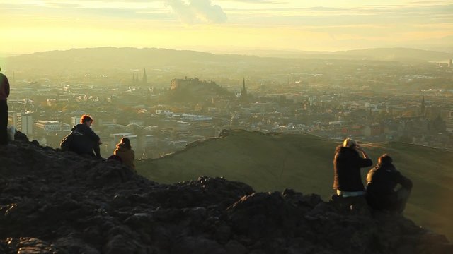 Tourists take in a view of distant Edinburgh, Scotland from the top of the King's Seat hike at sunset.
