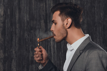 Handome brutal man in suit on the grey background lighting a cig