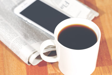 A black coffee with newspaper and a mobile phone. Image taken on a wooden table and has a vintage effect applied.