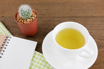 The cup of Japanese green tea and little cactus in plant pot and small note book with green cotton fabric on wooden planks.