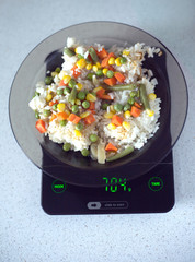 Translucent white plate with rice and vegetables is at home kitchen electronics scales to count calories in food. Top view photo closeup