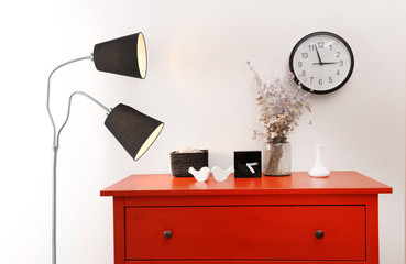 Room interior with red wooden commode, lamp and clock on light wall background