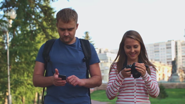 Young couple walking in the park: woman taking pictures with digital camera while her boyfriend using smartphone