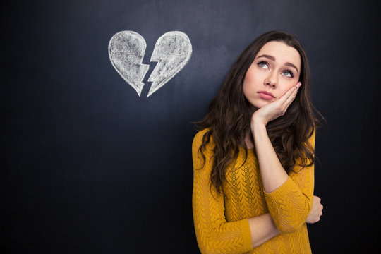 Upset woman thinking over chalkboard background with drawn broken heart