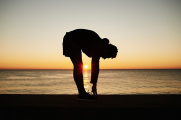 Young muscular build man doing stretching exercise on the beautiful sea background during sunset, attractive athlete doing workout outdoors at sunrise, fitness and healthy lifestyle concept.