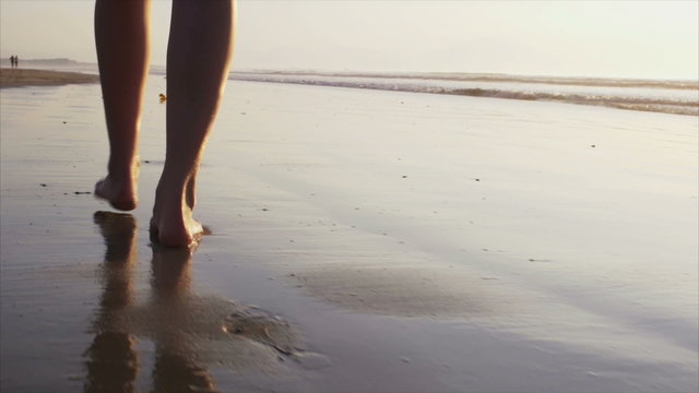 Slow motion video of woman walking on wet sea shore. Low section of female with bare feet on sand during sunset. Tracking shot of woman spending leisure time on beach.
